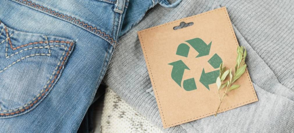 Sustainable still life with Blue denim jeans, gray pullover and craft paper card with Recycling symbol. Second hand apparel idea. Circular fashion, donation, charity concept. Top view