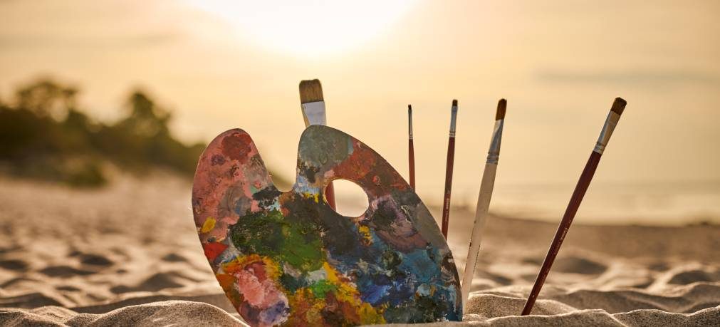 Painter palette and brushes rest on sandy beach, capturing serene ambiance of sunset. Scene symbolizes creativity in nature, evoking tranquility and artistic inspiration